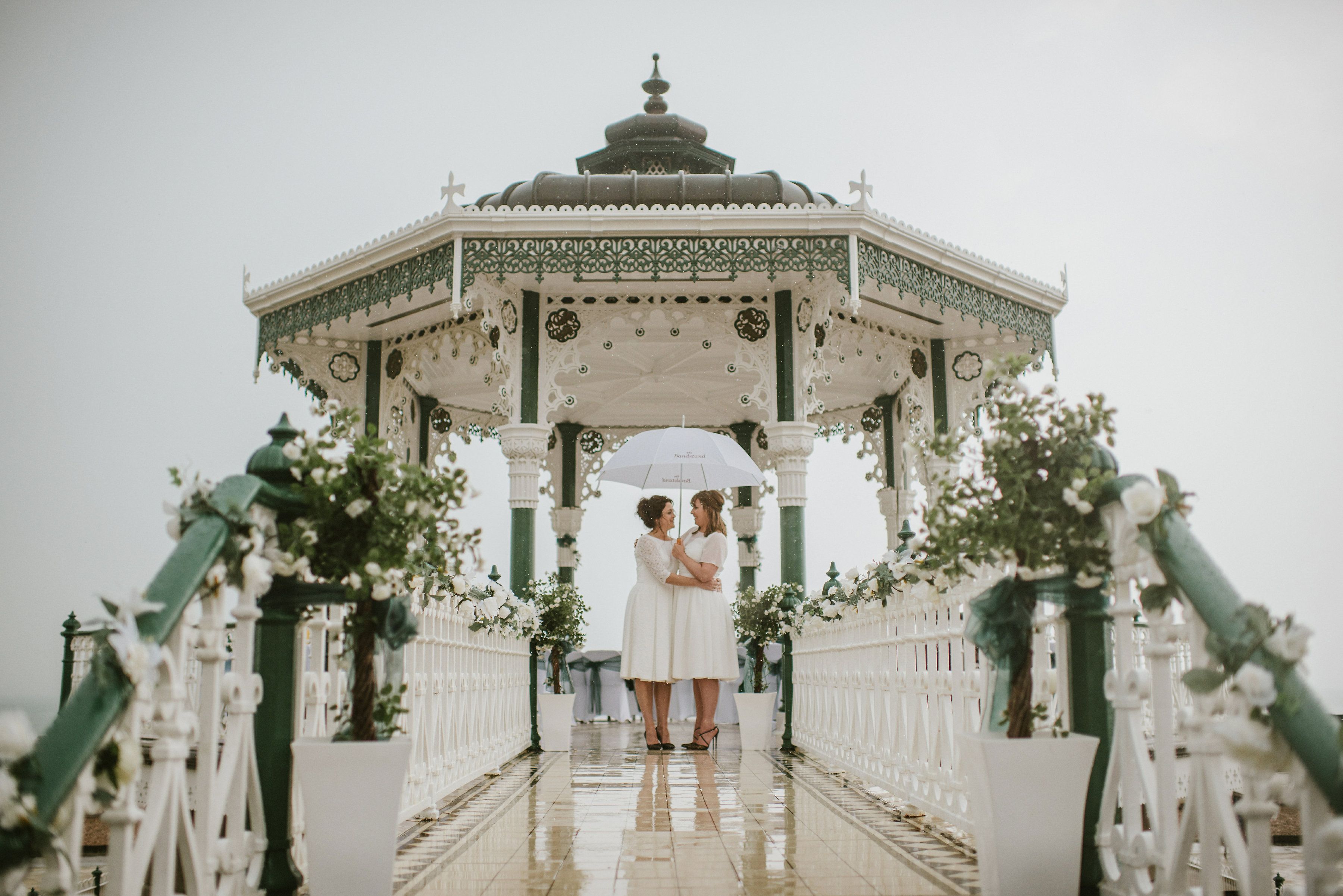 2 brides holding an umbrella, under a bandstand with lots of flowers