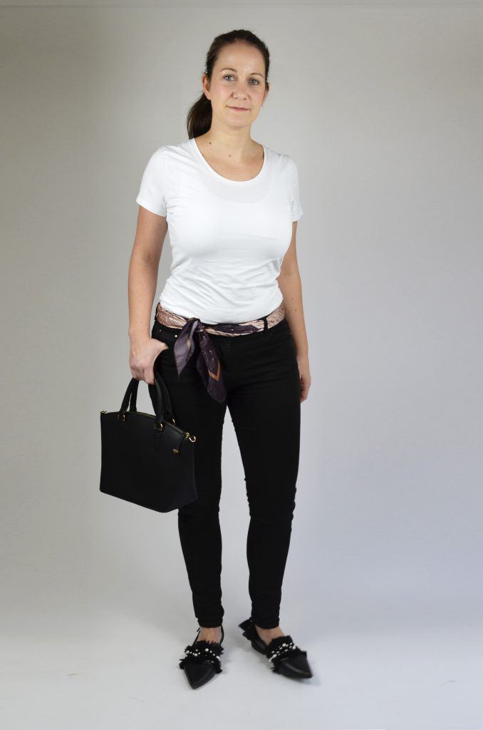 A white female model wearing black jeans and white t shirt holding a black bag and a Dara Ford scarf tied as a belt