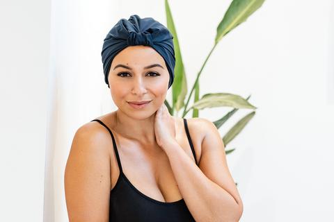 Woman wearing a navy blue silk headwrap and smiling She is standing in front of a green plant