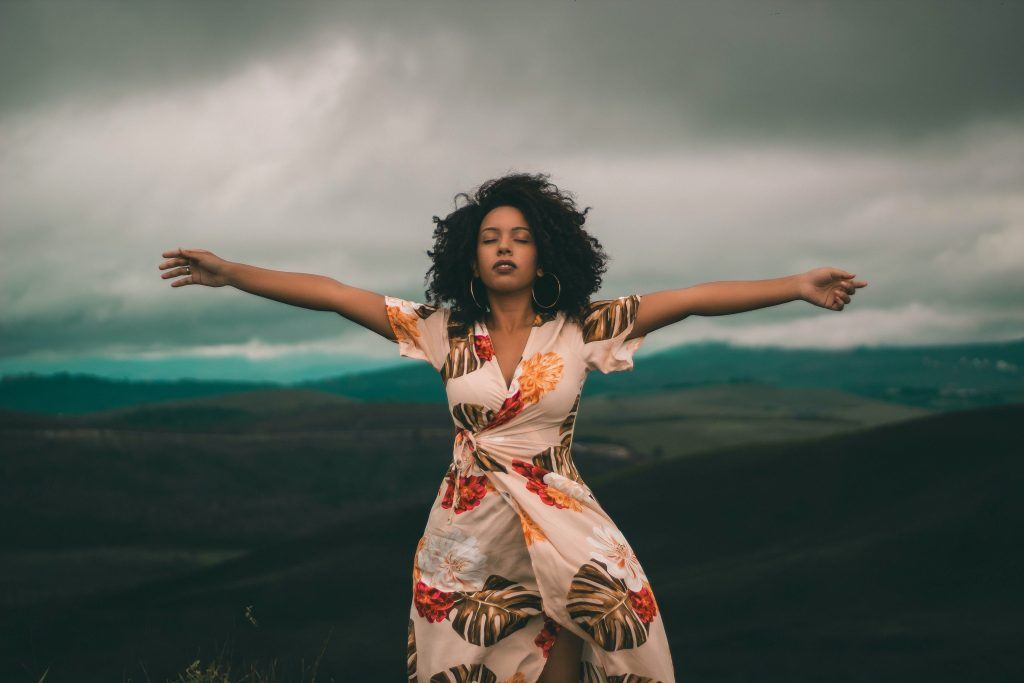 Black woman wearing a cream wrap dress with colourful leaf print arms spread wide and eyes closed Standing in front of a stormy landscape
