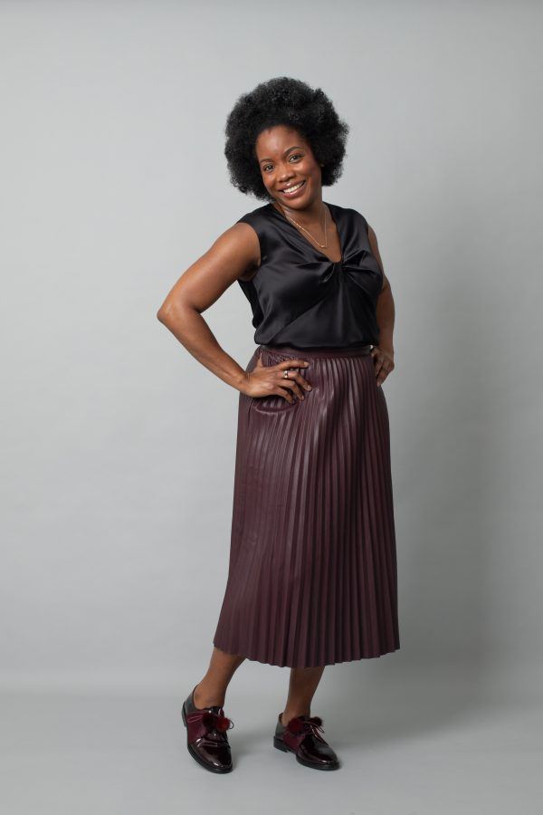 Full body image of black woman modelling black silk camisole with brown pleated faux leather skirt and burgundy brogues. Model is facing slightly to the side with her hands on her hips and smiling.