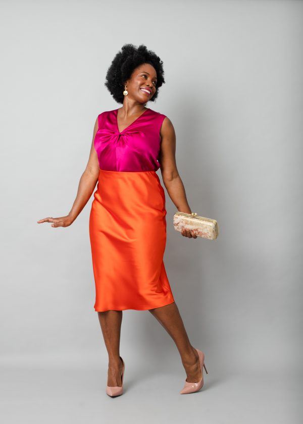 Party outfit modelled by black woman wearing pink silk blouse, orange satin skirt with high heels and carrying a gold clamshell clutch purse.