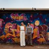 White Bride groom standing in front of a graffiti wall mural the bride is wearing a Dara Ford cropped blouse and trousers