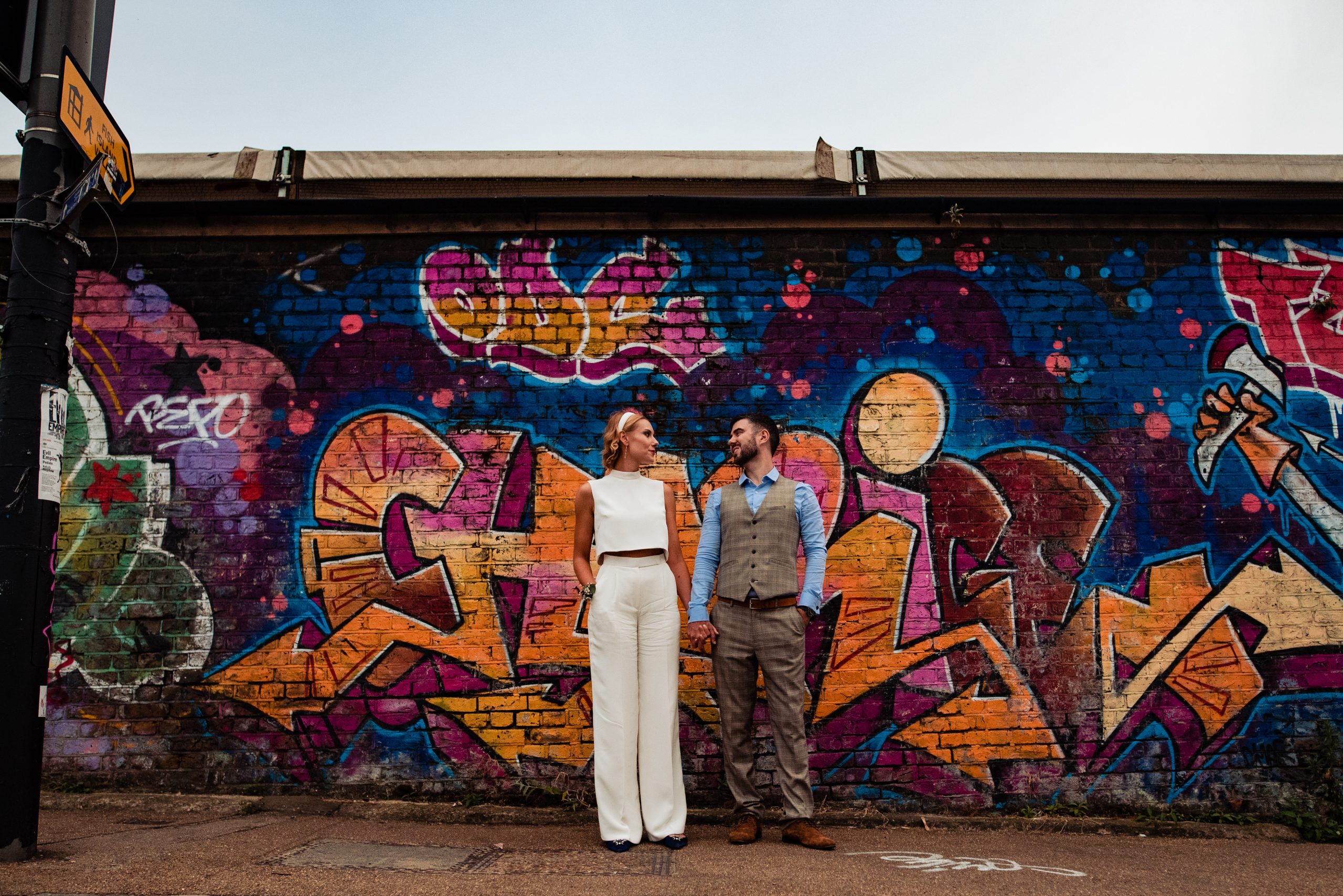 White Bride & groom standing in front of a graffiti wall mural, the bride is wearing a Dara Ford cropped blouse and trousers