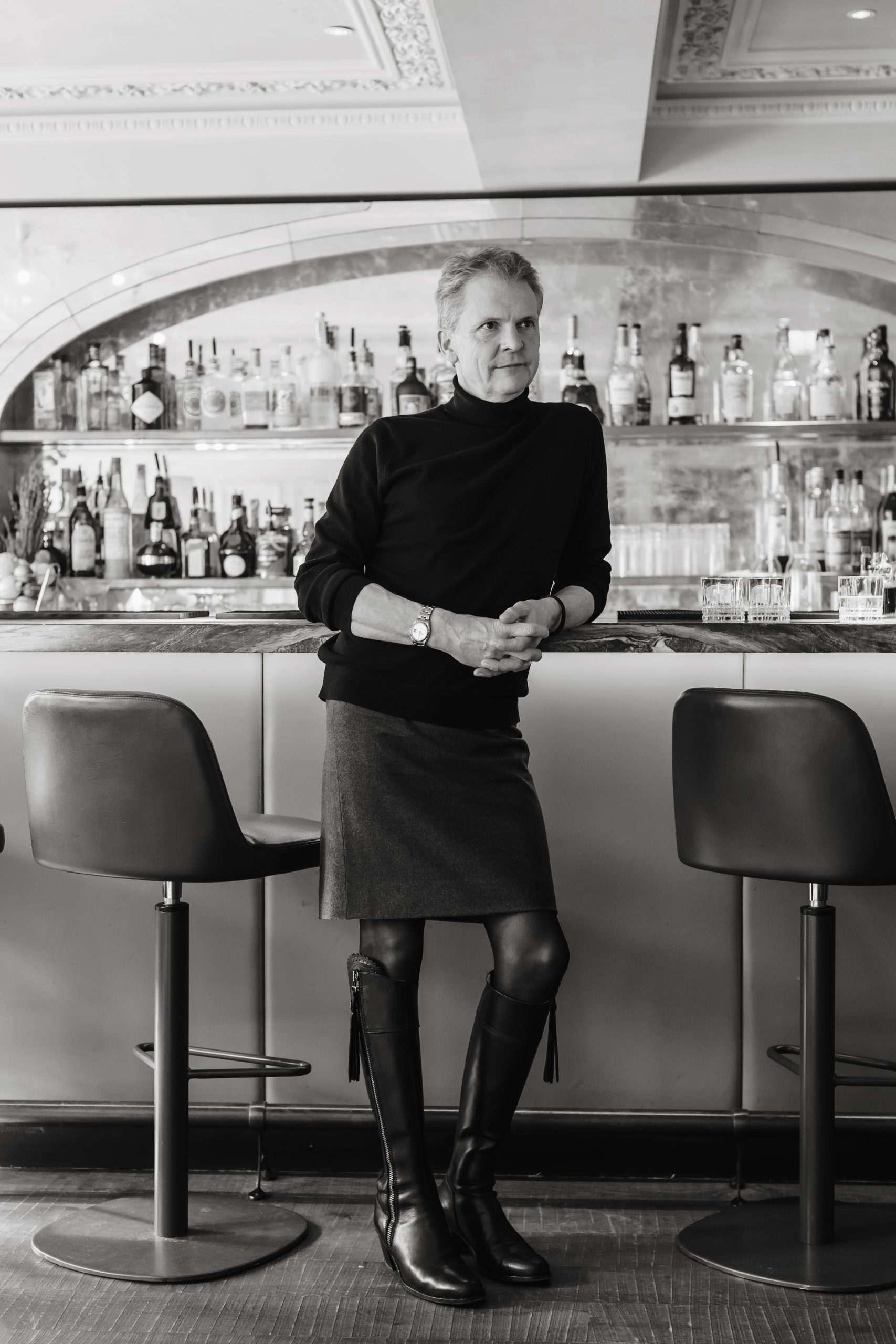 Man in his late 50s to 60s leaning against a bar and wearing a roll neck jumper with a tailored skirt, black tights and leather knee boots.