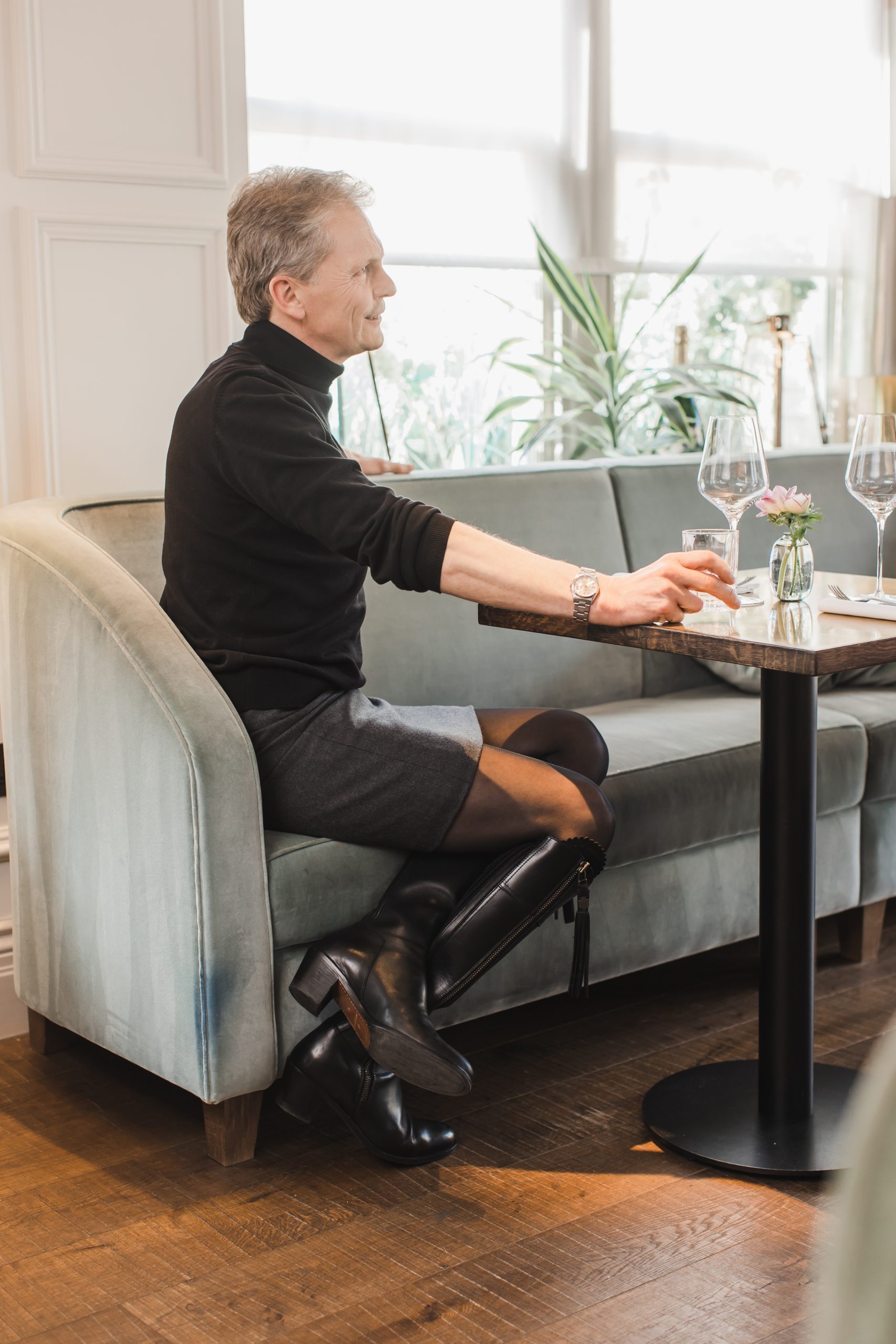Grey haired man sitting at a restaurant table wearing a tailored men's skirt, black turtleneck sweater and black boots.