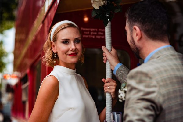 Bride and groom on red London bus. Bride is wearing sleeveless silk cropped blouse with stand collar and a matching Alice band.