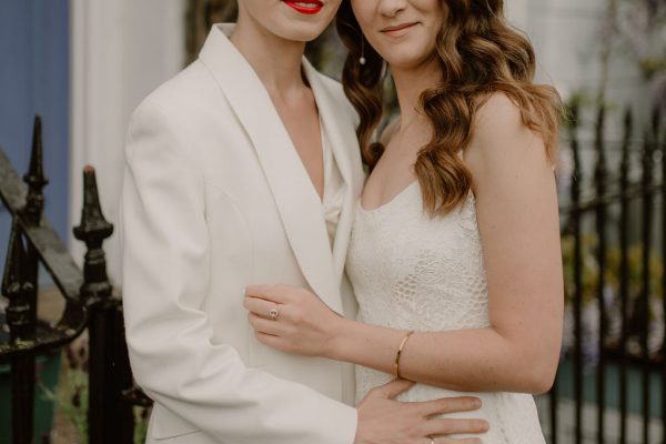 2 female models embracing, one wears a bridal trouser suit, the other a lace wedding dress in ivory