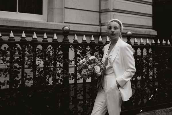 Black and white image of a bride in a tailored wedding suit with a veiled headband and heels, holding a bouquet