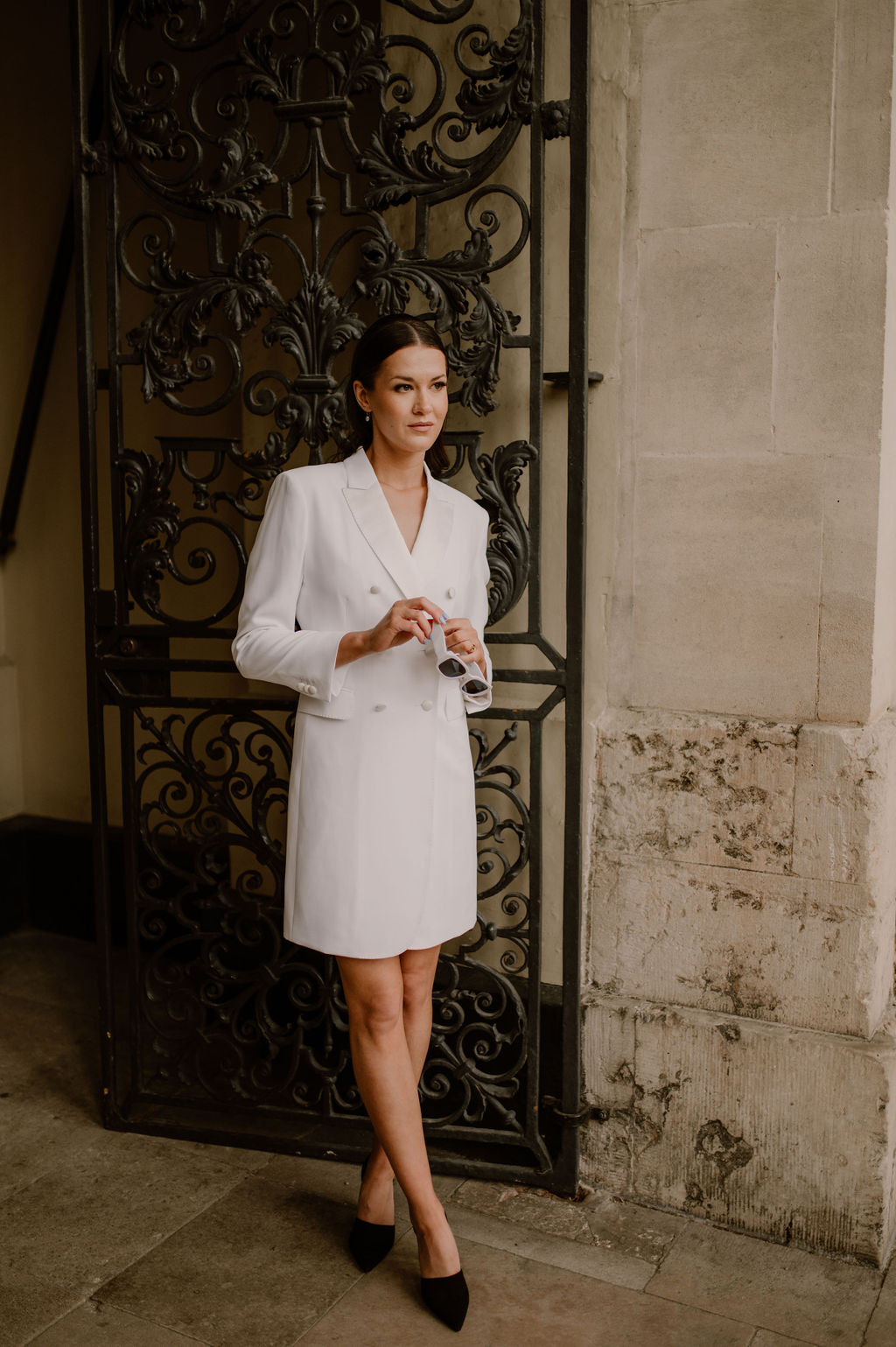 Brunette model stands outside an old iron gated building, wearing a blazer dress in ivory, holding a pair of white sunglasses